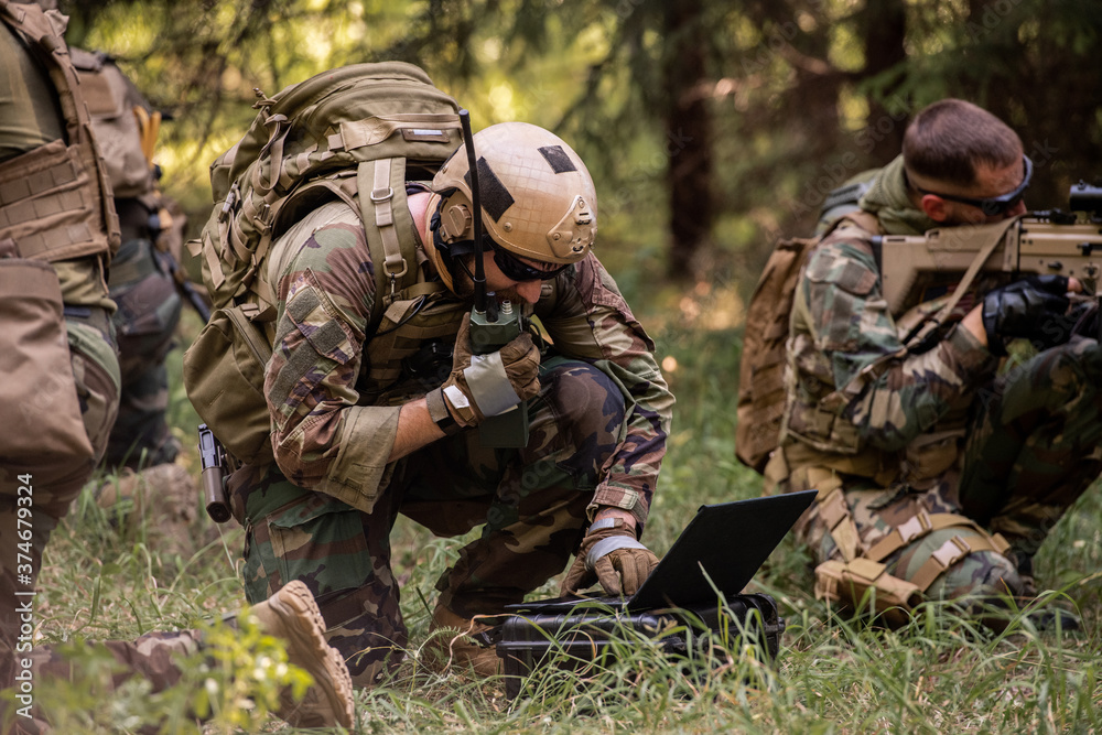 Busy soldier with backpack using military laptop while passing information through radio device in forest - obrazy, fototapety, plakaty 