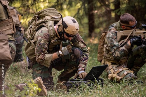 Busy soldier with backpack using military laptop while passing information through radio device in forest