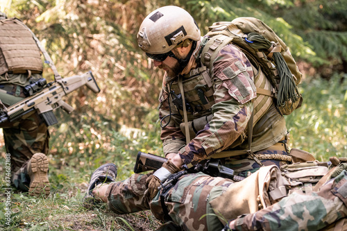 Konstfotografi Man in helmet and sunglasses examining leg of wounded soldier on battlefield