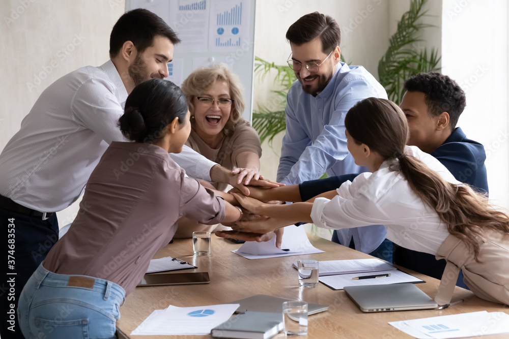 Overjoyed middle aged female team leader joining hands together with laughing young multiracial diverse colleagues over table, celebrating shared success or showing power in union gesture in office. Overjoyed middle aged female team leader joining hands together with laughing young multiracial diverse colleagues over table, celebrating shared success or showing power in union gesture in office.
