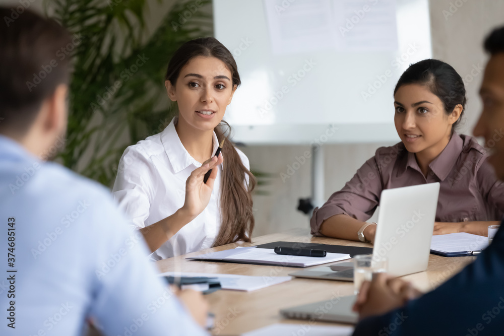 Confident young arab ethnicity woman sitting at table with diverse colleagues, explaining marketing strategy or developing project ideas at brainstorming meeting, office communication concept. Confident young arab ethnicity woman sitting at table with diverse colleagues, explaining marketing strategy or developing project ideas at brainstorming meeting, office communication concept.
