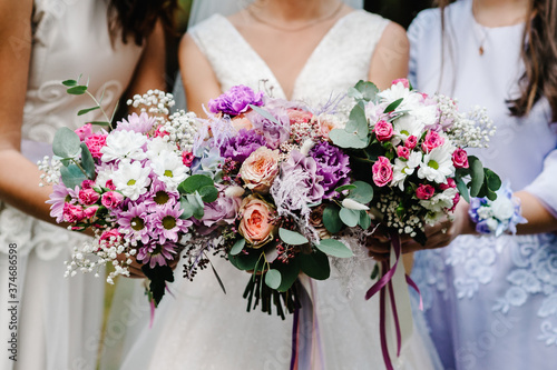 The bride and bridesmaids in an elegant dress is standing and holding hand bouquets of pastel pink flowers and greens with ribbon at nature. Young beautiful girls holds a wedding bouquet outdoors.