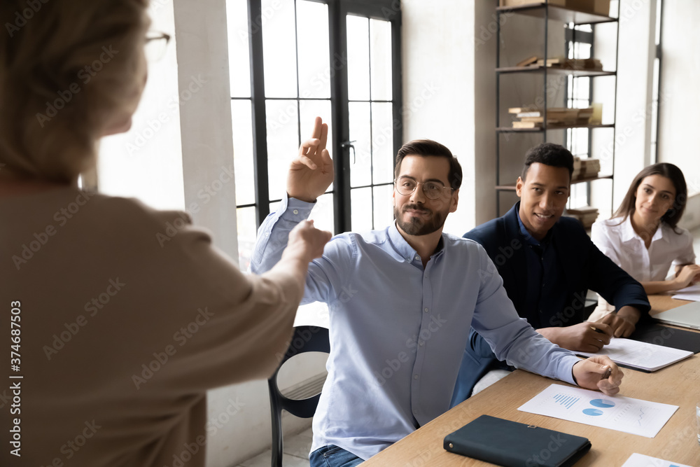 Initiative interested young businessman in eyeglasses raising hand, asking questions about marketing strategy to older senior female leader at presentation meeting with diverse colleagues in office. Initiative interested young businessman in eyeglasses raising hand, asking questions about marketing strategy to older senior female leader at presentation meeting with diverse colleagues in office.