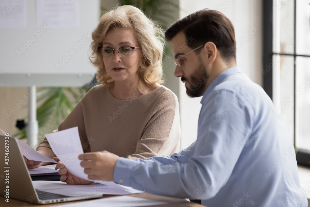 Confident focused middle aged 50s businesswoman in eyeglasses discussing marketing research results or company sales statistics paper reports with skilled young male employee at office meeting. Confident focused middle aged 50s businesswoman in eyeglasses discussing marketing research results or company sales statistics paper reports with skilled young male employee at office meeting.