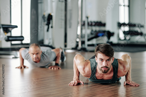 Handsome young fitness trainer showing half cobra push ups to mature client in gym