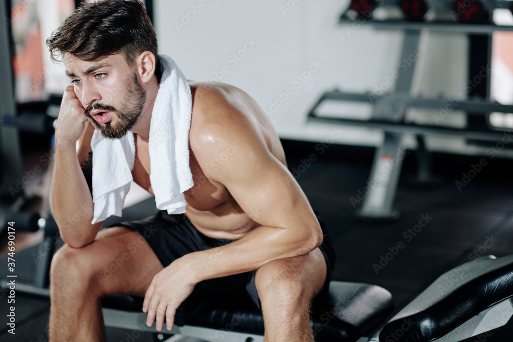 Exhausted handsome fit young man with towel sitting on bench and resting after training in gym Exhausted handsome fit young man with towel sitting on bench and resting after training in gym