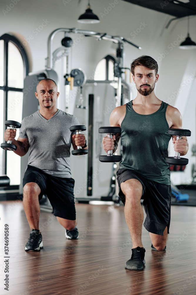 Mature mixed-race man and his fitness trainer doinglunges with heavy dumbbells in hands