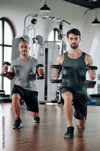 Mature mixed-race man and his fitness trainer doinglunges with heavy dumbbells in hands