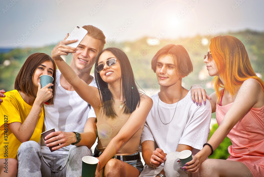 Obraz premium Friends drinking coffee on the background of the city. Boys and girls are smiling. Teenagers sitting on the curb on a sunny day in the square. Young people taking selfies.