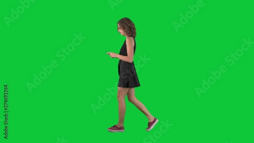 Young stylish woman walking and messaging using cell phone. Full body side view on green screen chroma key background. 