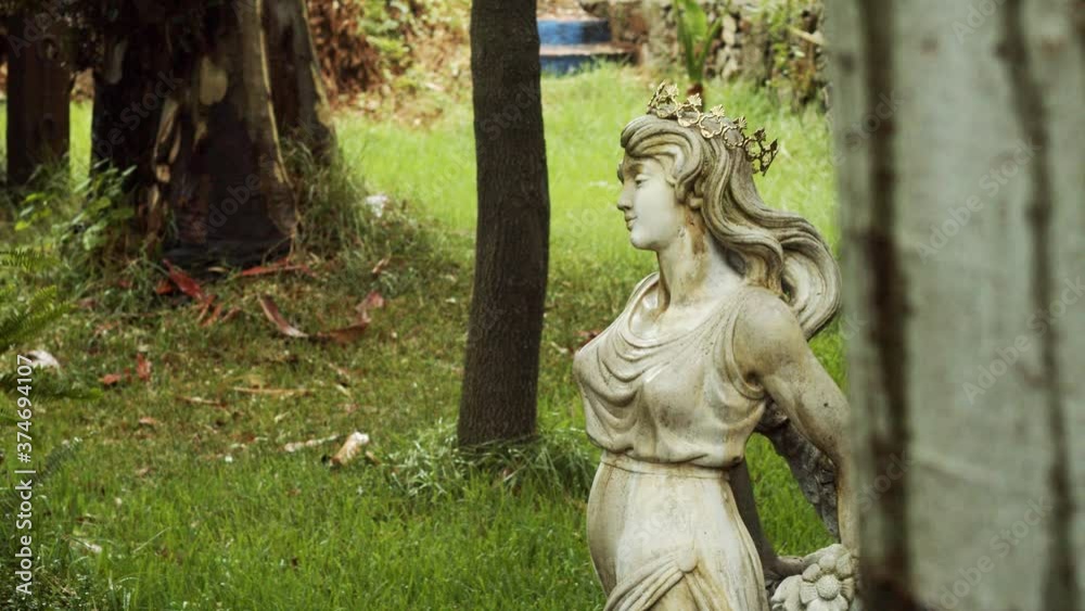 Static shot for a female white statue with a golden crown and long hair standing between trees in the middle of a garden with green grass. Static shot for a female white statue with a golden crown and long hair standing between trees in the middle of a garden with green grass.
