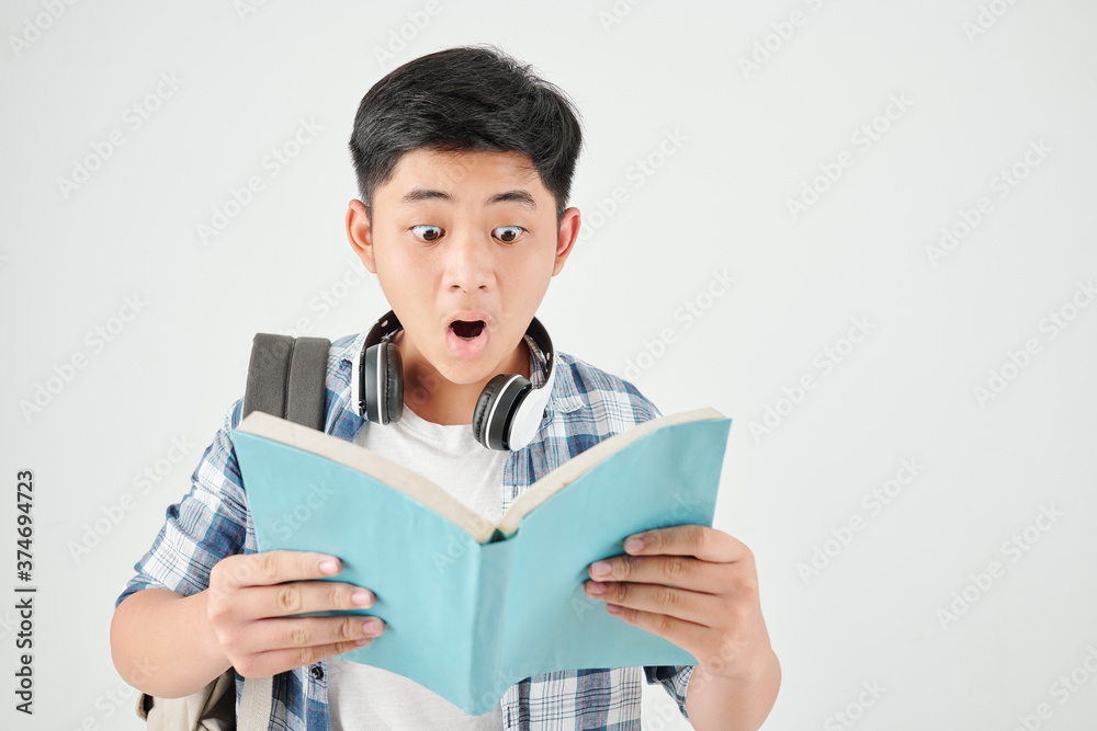 Studio portrait of shocked excited schoolboy with backpack reading text in book Studio portrait of shocked excited schoolboy with backpack reading text in book