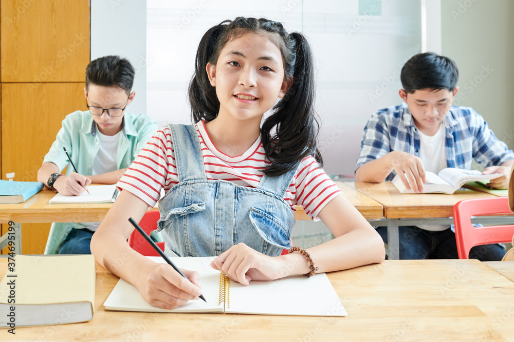 Smiling Asian schoolgirl writing essay in textbook during English class Smiling Asian schoolgirl writing essay in textbook during English class