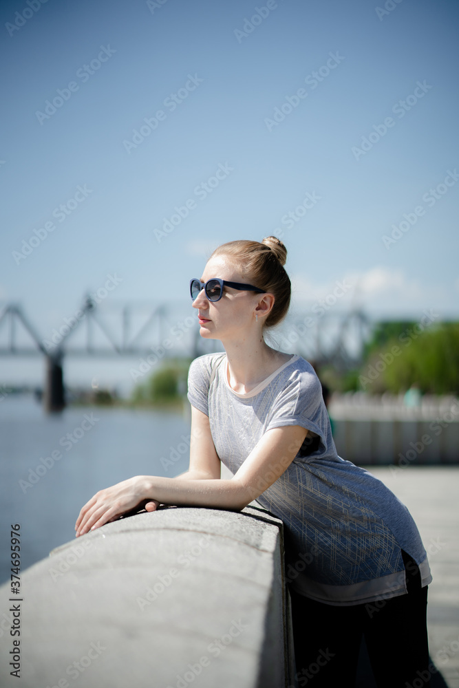 A young woman in sunglasses stands on the embankment of the river against the background of a bridge on a sunny summer day. Reflections alone, vacation in an urban environment or summer walks concept