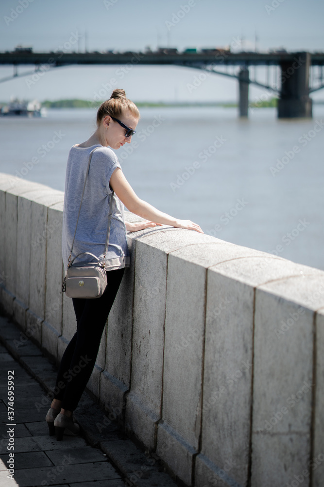 A young woman in sunglasses stands on the embankment of the river against the background of a bridge on a sunny summer day. Reflections alone, vacation in an urban environment or summer walks concept.
