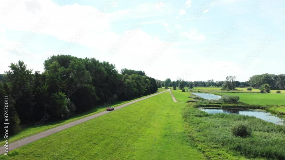 Red Cabriolet Car Driving And Travelling On The Road With Green Hills At Daytime In Zwolle, Netherlands. - aerial drone Red Cabriolet Car Driving And Travelling On The Road With Green Hills At Daytime In Zwolle, Netherlands. - aerial drone