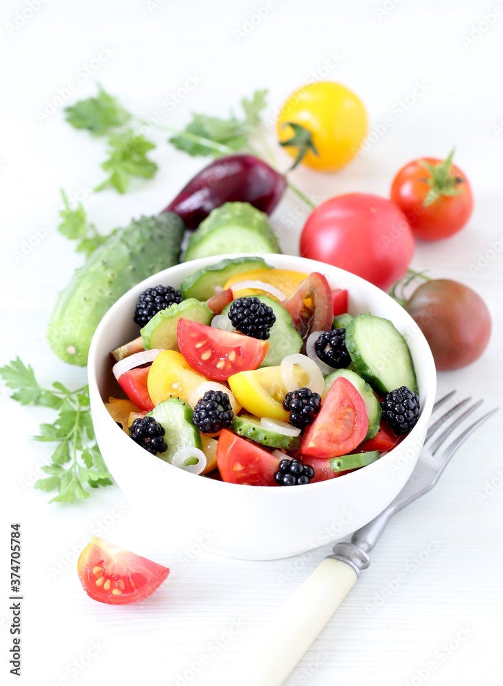 Vegetable berry salad of fresh yellow, red, black tomatoes, cucumbers, onions and blackberries in a bowl on a white background