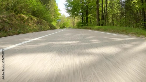 POV, LENS FLARE: Speeding down an empty asphalt road winding through a lush forest on a sunny spring day. Carefree joyride through the idyllic green Slovenian countryside in a powerful supercar. photo