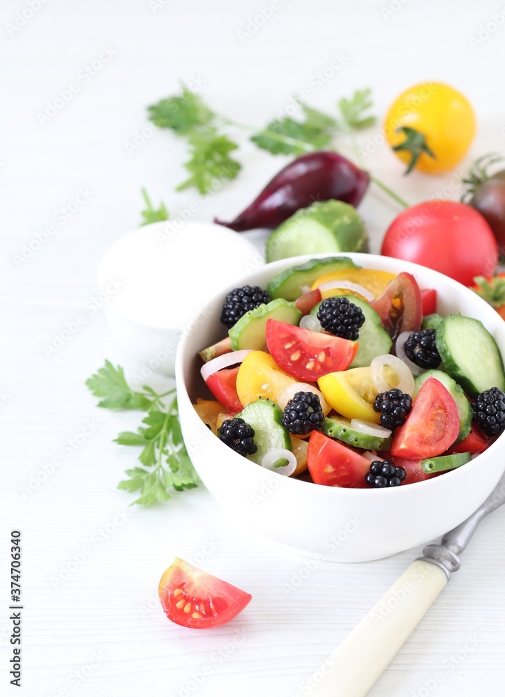Vegetable berry salad of fresh yellow, red, black tomatoes, cucumbers, onions and blackberries in a bowl on a light background
