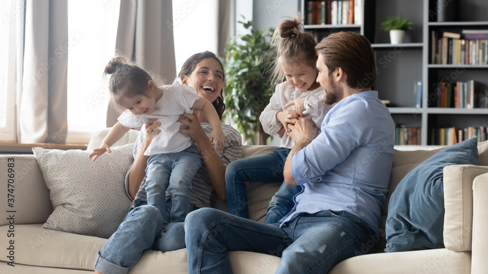 Joyful carefree parents playing with adorable little kids, resting on comfortable couch together at home. Positive happy different generations family having fun with preschool baby daughters indoors.