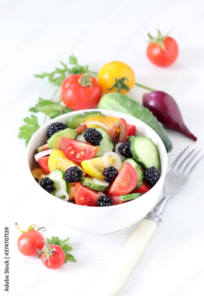 Vegetable berry salad of fresh yellow, red, black tomatoes, cucumbers, onions and blackberries in a bowl on a white background