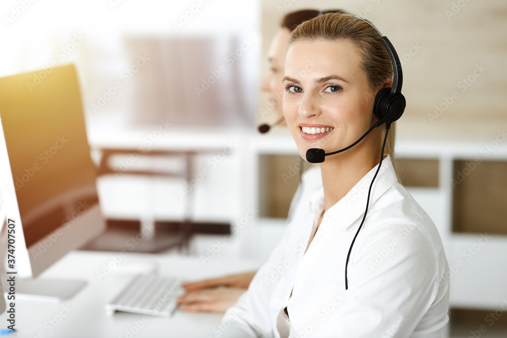 Blond female customer service representative is consulting clients online using headset in sunny office. Call center concept Blond female customer service representative is consulting clients online using headset in sunny office. Call center concept