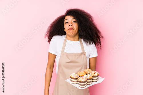 Young afro pastry maker woman holding a cupcakes isolatedYoung afro baker woman shrugs shoulders and open eyes confused.