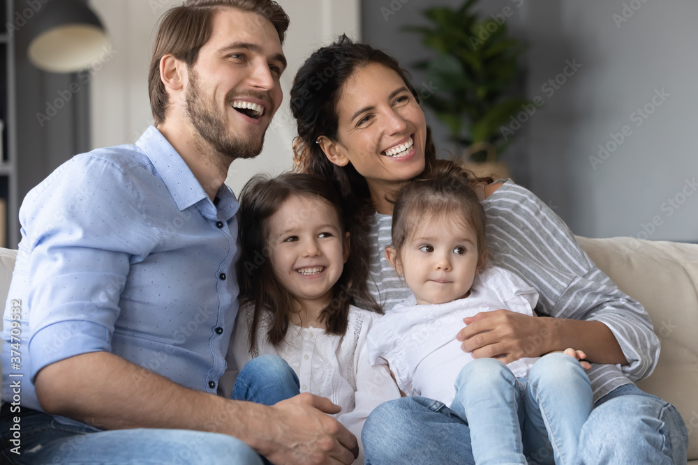 Smiling young married couple relaxing on sofa with adorable little children daughters, enjoying watching TV program, kids show or cartoons together at home, happy family hobby weekend pastime. Smiling young married couple relaxing on sofa with adorable little children daughters, enjoying watching TV program, kids show or cartoons together at home, happy family hobby weekend pastime.