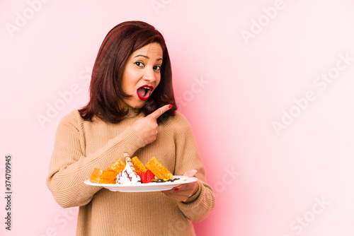 Middle age latin woman holding a waffle isolated pointing to the side