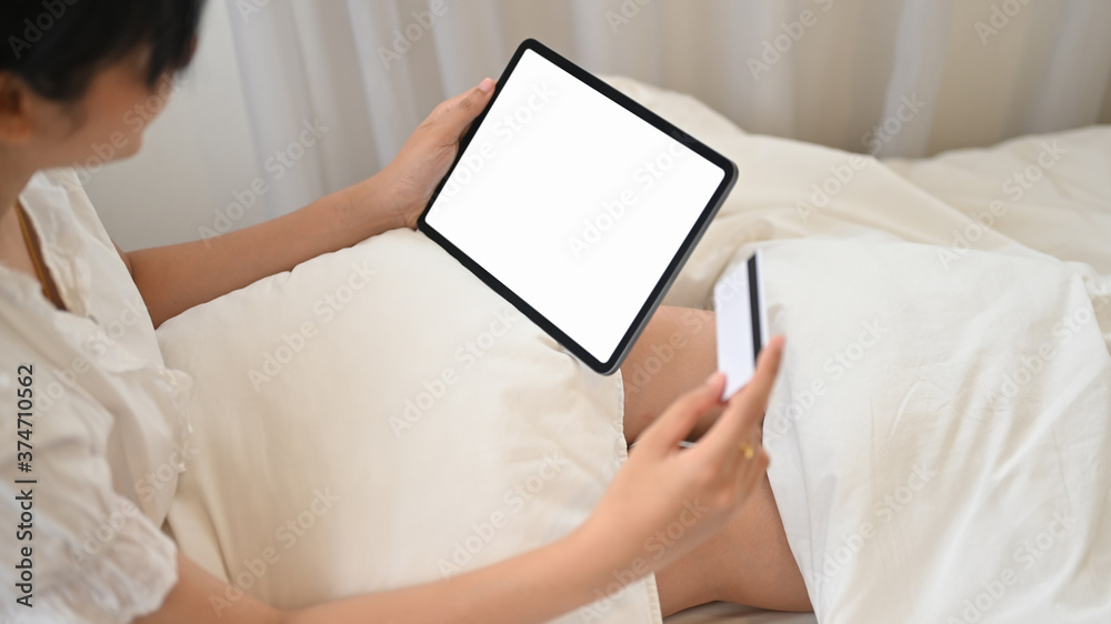A woman in white pajamas is using an empty screen computer tablet and holding a credit card in hand. A woman in white pajamas is using an empty screen computer tablet and holding a credit card in hand.