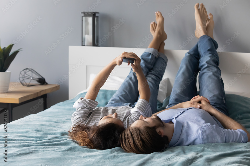 Relaxed young couple lying on comfortable bed, posing for selfie shot on smartphone. Happy millennial family of two resting in bedroom, recording stories for social network, shopping or web surfing.