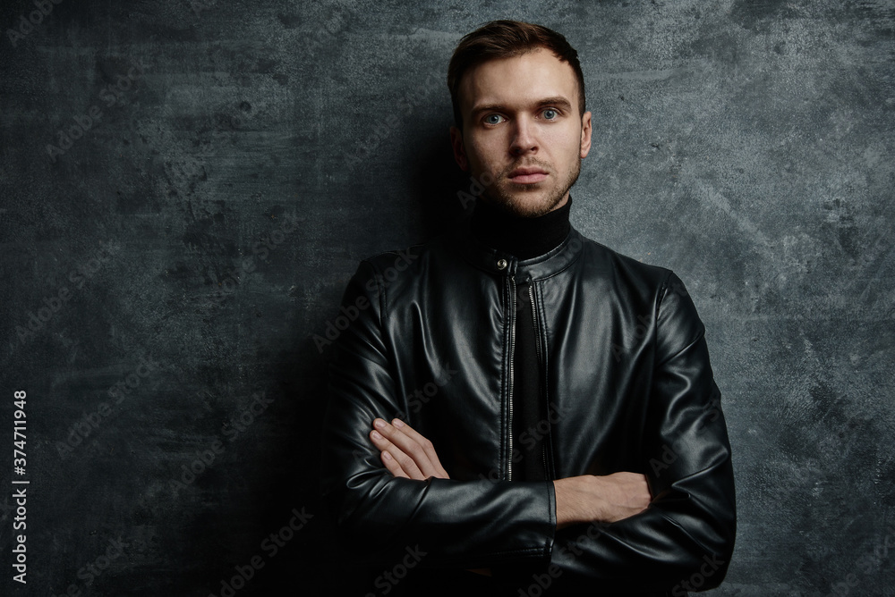 Caucasian unshaven sexy male model with brutal serious face in black leather jacket with crossed arms looking at camera on dark background isolated