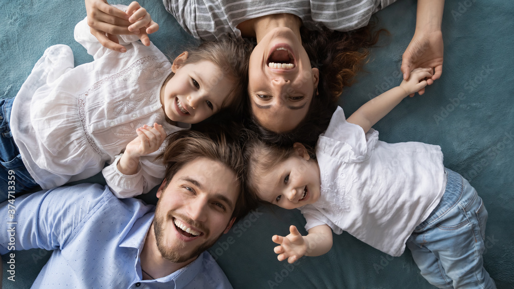 Top above view happy young parents relaxing on bed with joyful adorable little baby girls at home. Portrait of energetic emotional couple having fun with small kids indoors, looking together at camera Top above view happy young parents relaxing on bed with joyful adorable little baby girls at home. Portrait of energetic emotional couple having fun with small kids indoors, looking together at camera