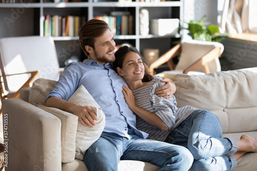 Romantic young bonding couple embracing on sofa, looking away, dreaming visualizing good future together at home. Happy man cuddling smiling woman, watching television programs, weekend pastime.