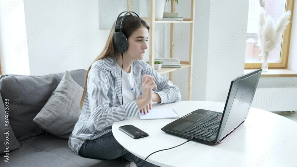 Serious girl student wears headphones, studies with internet teacher, learns language. focused young woman talks to tutor, looks at laptop, writes notes. online education, video call tutoring concept Serious girl student wears headphones, studies with internet teacher, learns language. focused young woman talks to tutor, looks at laptop, writes notes. online education, video call tutoring concept