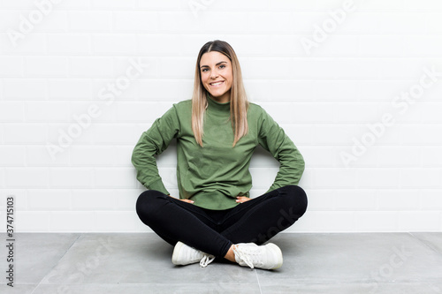 Young caucasian woman sitting on the floor confident keeping hands on hips.