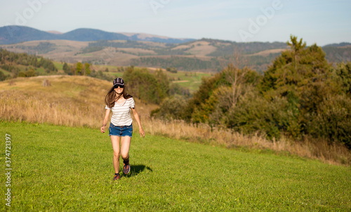 Carefree happy woman in sunglasses, cap and jeans shorts walk on green grass meadow on top of mountain enjoying nature. Freedom