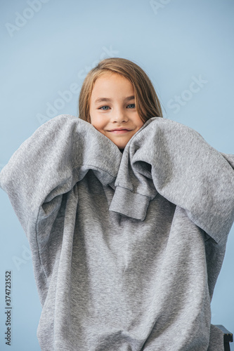 Smiling little girl in a huge oversized grey longsleeve  playing cute  touching chin with hand inside overly long sleeves. Over blue background.