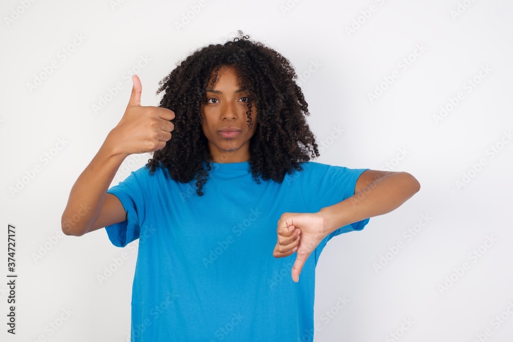 Young african woman with curly hair wearing casual blue shirt over white background showing thumbs up and thumbs down, difficult choose concept Young african woman with curly hair wearing casual blue shirt over white background showing thumbs up and thumbs down, difficult choose concept