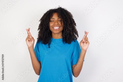 Positive Young african woman with curly hair wearing casual blue shirt over white background has big hope, crosses fingers, believes in good fortune, smiles broadly. People and wish concept © Roquillo