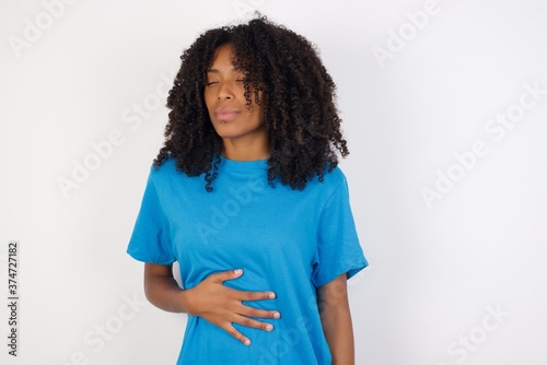 Young african woman with curly hair wearing casual blue shirt over white background touches tummy, smiles gently, eating and satisfaction concept.