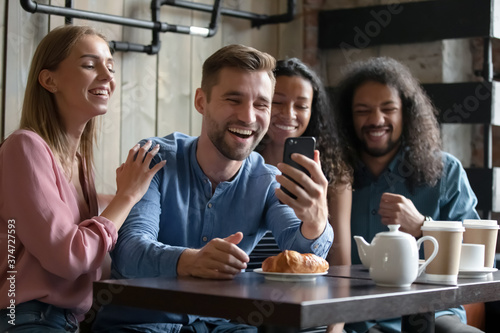 Overjoyed diverse young people posing for photo in cafe together, smiling excited man holding smartphone, taking selfie or recording video for social network, having fun with friends
