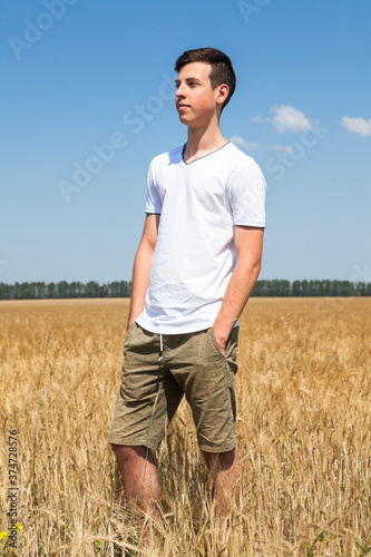Full length portrait of teen age boy standing on agricultural field with ripe wheat, yellow meadow and blue sky, hands are in pockets of shorts pants © Kekyalyaynen