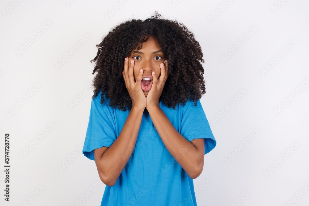 Stupefied Young african woman with curly hair wearing casual blue shirt, expresses excitement and thrill, keeps jaw dropped, hands on cheeks, has eyes popped out Stupefied Young african woman with curly hair wearing casual blue shirt, expresses excitement and thrill, keeps jaw dropped, hands on cheeks, has eyes popped out