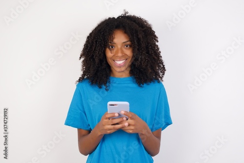Photo of Young african woman enjoys distant communication, uses mobile phone, surfs fast unlimited internet, has pleasant smile, dressed in blue t-shirt, makes shopping online, isolated on white