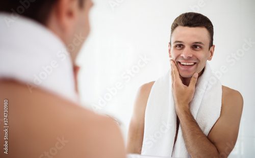 Handsome young man shaving his beard in the bathroom. Portrait of a stylish naked bearded man examining his face in-home mirror.