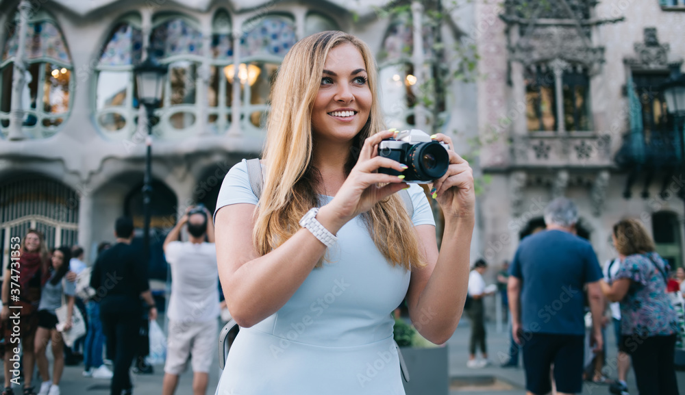 Pretty Caucasian tourist with retro camera smiling at Spanish street with architecture building of Antonio Gaudi design on background, cheerful woman with vintage equipment exploring Barcelona