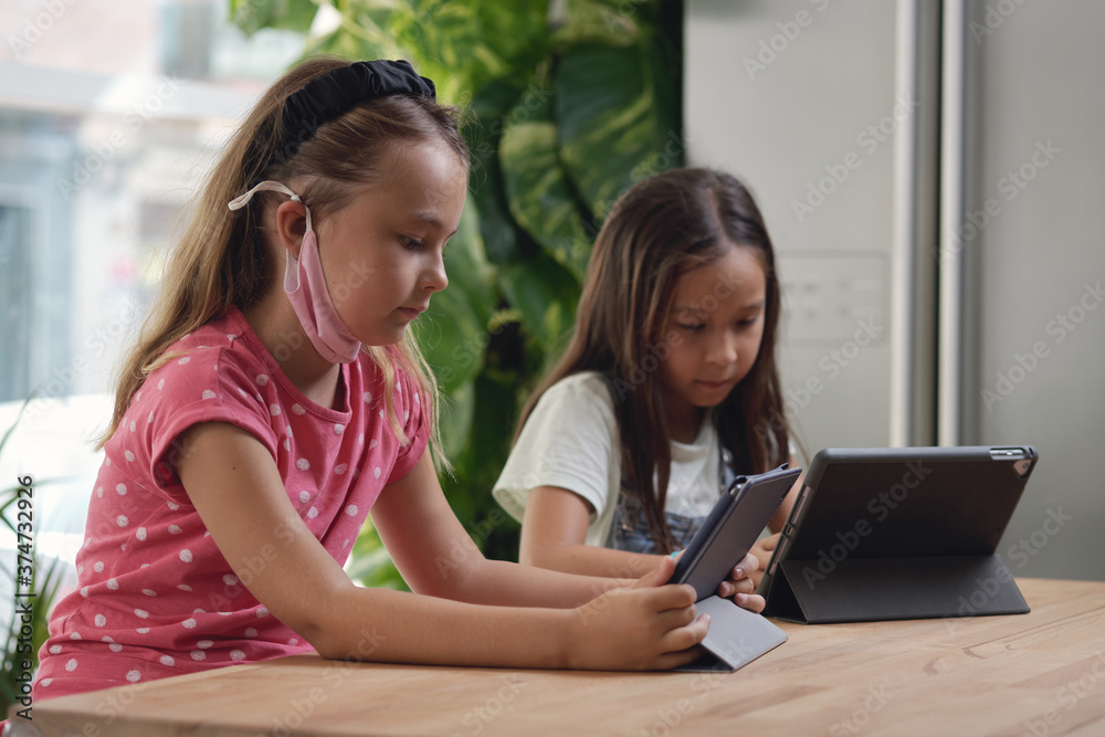 Two multi racial beautiful school age 9s girls sit at desk with portable computer. Using modern device ignoring each other, studying distantly by wireless gadget, new generation tablet overuse concept Two multi racial beautiful school age 9s girls sit at desk with portable computer. Using modern device ignoring each other, studying distantly by wireless gadget, new generation tablet overuse concept
