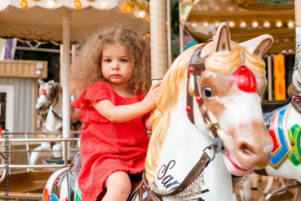 A little curly girl in a red dress sits on a horse in an amusement park on a carousel