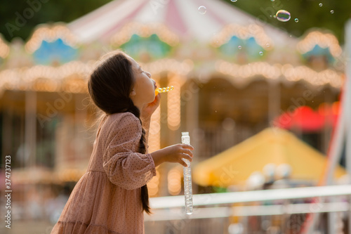 Teenage girl with two pigtails launches bubbles in amusement park near carousel and rejoices